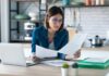 young woman working with computer while consulting some invoices and documents in the kitchen