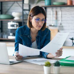 young woman working with computer while consulting some invoices and documents in the kitchen